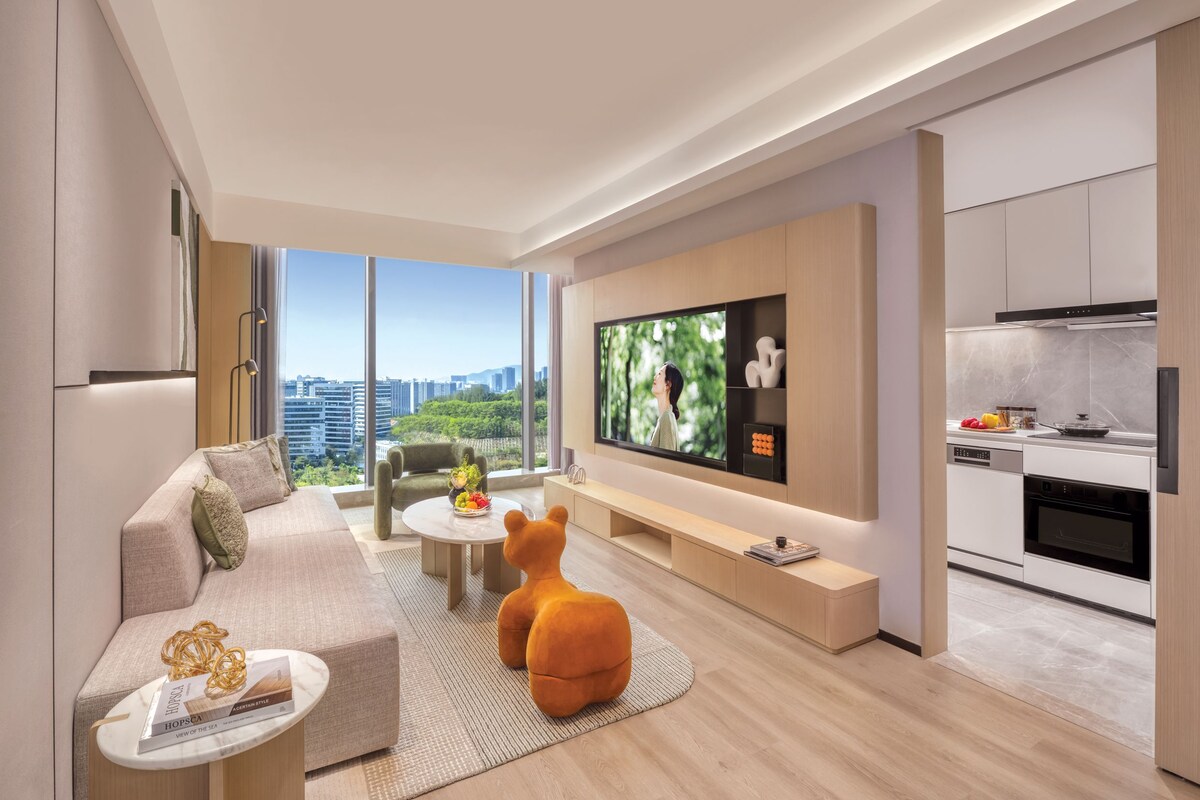 Una sala moderna con un sofá, una mesa de centro y un pouf con forma de oso. Un televisor de pantalla plana está montado en la pared, y hay una zona de cocina con estufa y horno.