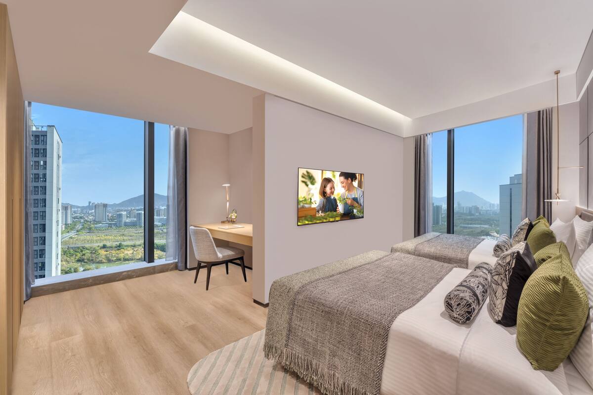 A modern hotel room with a large window, a desk, and a bed with decorative pillows.