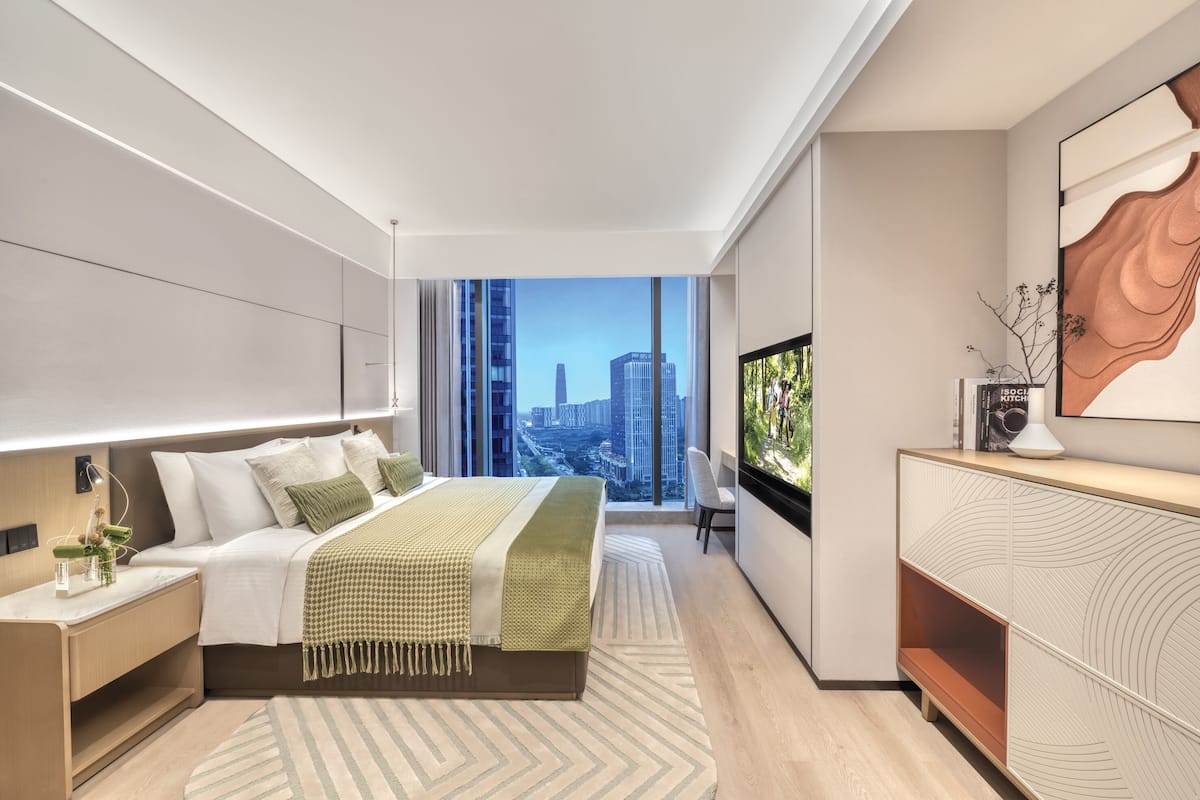 A modern hotel room with a large bed, a TV, a desk, and a view of the cityscape.