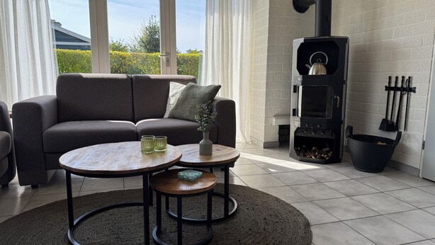 En moderne stue med sofa, to runde borde, en vase og en brændeovn.