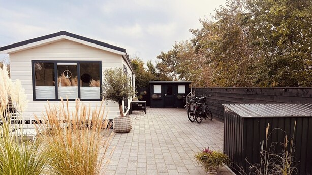 Ein modernes Mobilheim mit befestigter Auffahrt, einer kleinen Terrasse und einem Garten mit verschiedenen Pflanzen.
