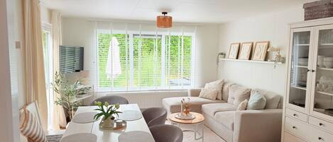 Ein modernes Wohnzimmer mit Essbereich, einer Sofagarnitur, einem Schrank mit Glastüren und einem großen Fenster mit Jalousien.