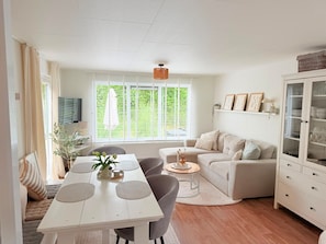 A modern living room with a dining area, a sofa set, a cabinet with glass doors, and a large window with blinds. - Chalet 229 (4 persons) (Petten)