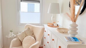 Ein Zimmer mit weißem Sessel, einer weißen Kommode mit Schubladen, einem kleinen Beistelltisch, einer Lampe und einem Fenster mit Jalousien.