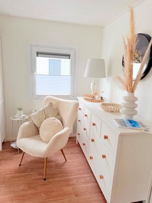 A room with a white chair, a white dresser with drawers, a small side table, a lamp, and a window with blinds. - Chalet 229 (4 persons) (Petten)