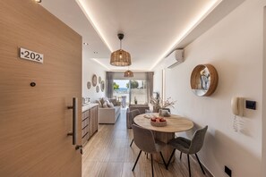 A modern hotel room with a wooden door, a number sign, a dining area with a round table, chairs, and a hanging light fixture. - 2 Bedroom Apartment In Fig Tree Bay (protaras)