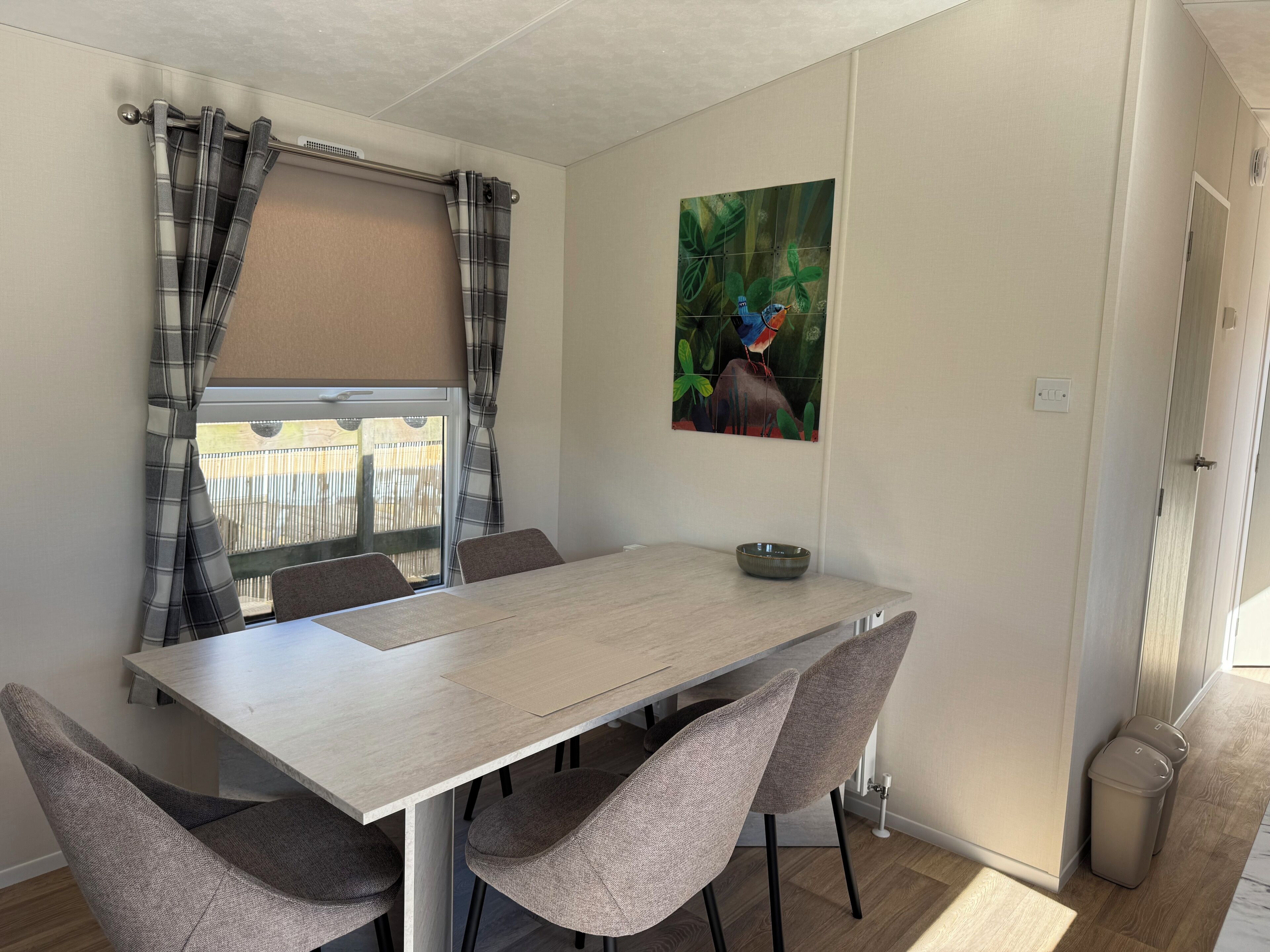 Een eethoek met een tafel, stoelen, een raam met gordijnen, een aan de muur hangend schilderij en een deur.