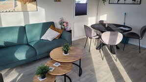 A living room with a teal sofa, a wooden coffee table, a dining table with chairs, a world map on the wall, and a window with blinds. - Chalet 34 (4 persons) (Petten)