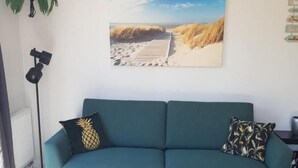 A teal sofa with a pineapple pillow, two round wooden coffee tables, a potted plant, and a beach scene painting on the wall. - Chalet 34 (4 persons) (Petten)