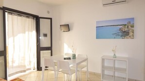 Una habitación con una cama, una mesa con sillas, un armario blanco pequeño, un cuadro de una escena costera y un equipo de aire acondicionado.
