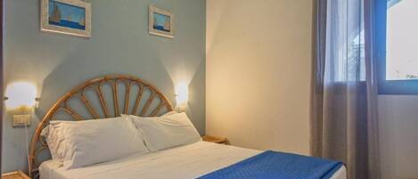 A bedroom with a bed, two wall-mounted lamps, a window with curtains, and two framed pictures on the wall.