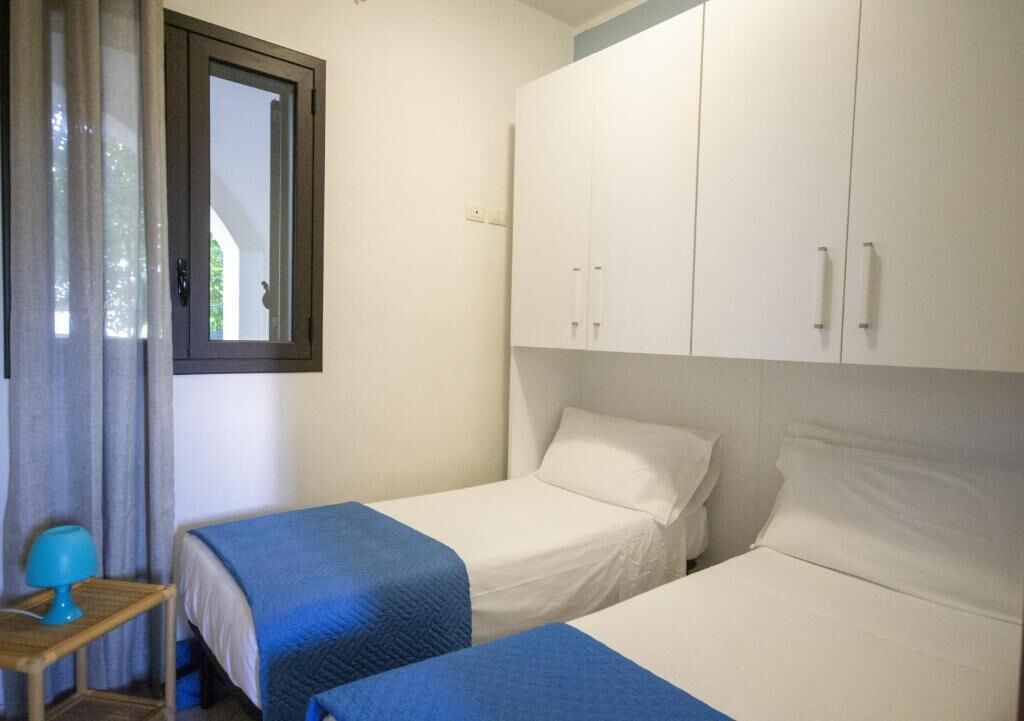 Un dormitorio con dos camas, una ventana con cortinas, una mesita de noche con lámpara y armarios blancos.