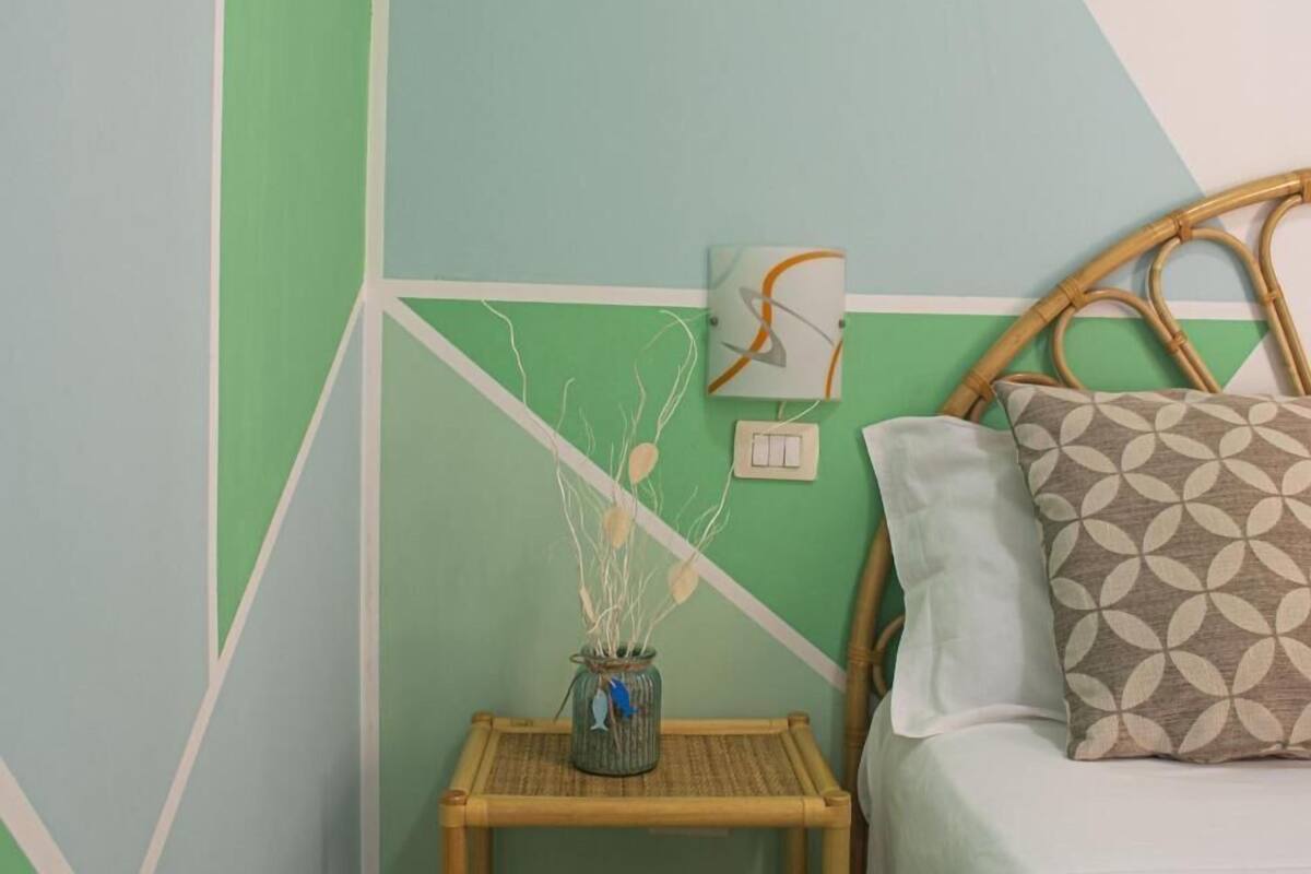 A room with a geometric-patterned wall, a bed with a patterned pillow, a small table with a vase, and a wall-mounted light switch.