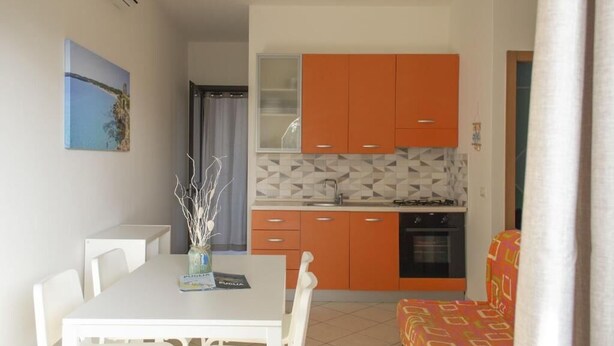 Cozinha compacta com armários laranja, bancada branca e um espaço de refeições com mesa e cadeiras brancas.
