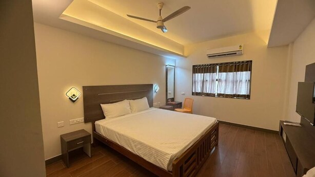 A bedroom with a bed, a nightstand, a ceiling fan, an air conditioning unit, and a window with curtains. - Sam's Court Elite Stay (Manachanallur)