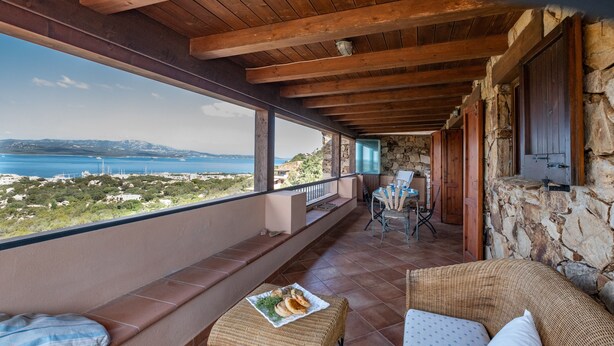 Een balkon met een stenen muur, houten plafond, rieten meubels en uitzicht op zee.