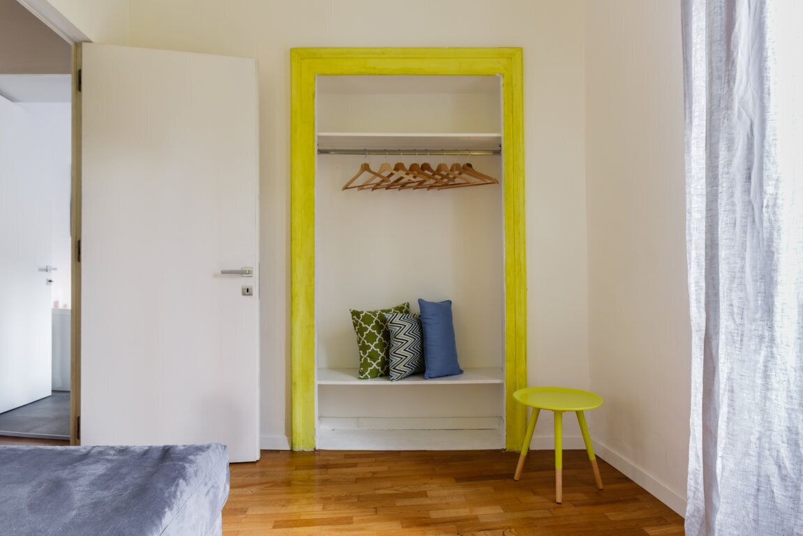 Una stanza con pavimento in legno, un letto, un armadio con cornice gialla, una panca con cuscini e un piccolo tavolo giallo.