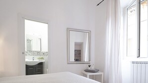 A bedroom with a bed, a bedside table, a mirror, a window with curtains, and a bathroom visible through an open door. - Santonofrio Silver - One Bedroom Apartment, Sleeps 4 (Rome)