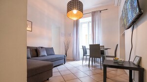 A living room with a sofa, a dining table with chairs, a hanging light fixture, and a TV mounted on the wall. - Santonofrio Silver - One Bedroom Apartment, Sleeps 4 (Rome)