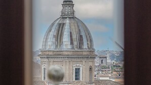 Vista de un edificio con cúpula y cruz en la parte superior, probablemente una iglesia o catedral, visible a través de una ventana con marco de madera y una planta en primer plano.