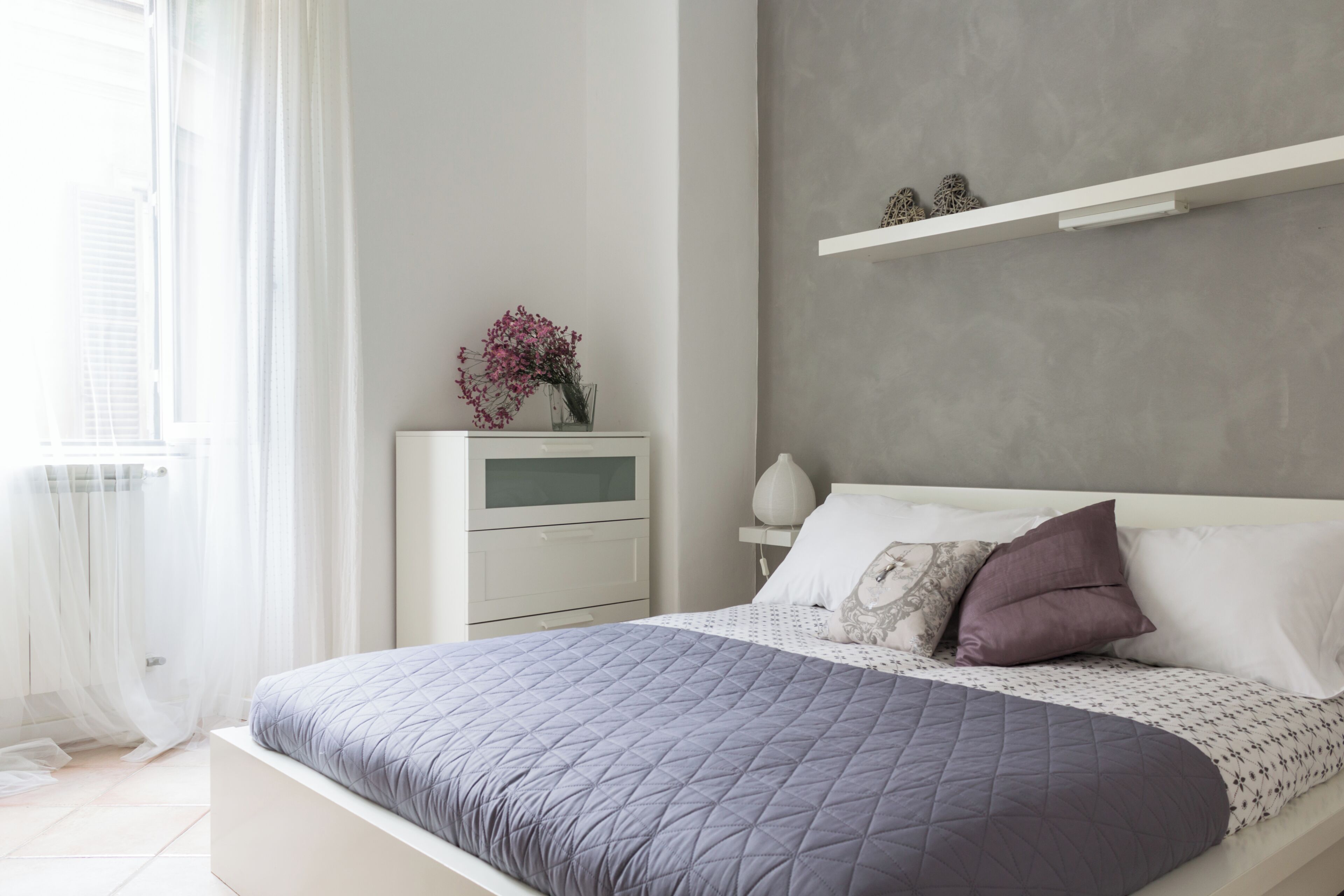 Een slaapkamer met een bed, nachtkastje en een raam met dunne gordijnen.