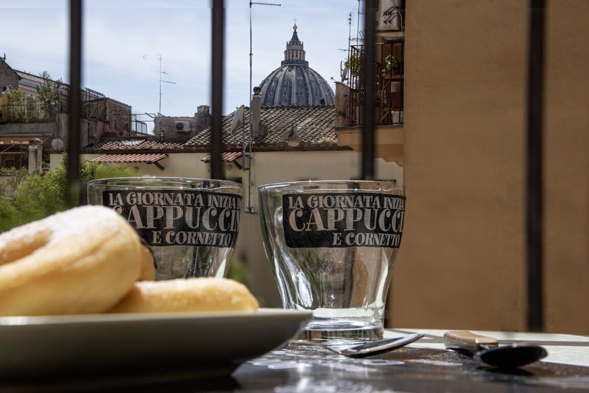 Una mesa con dos vasos etiquetados 'La Giornata Nella Cappuccio e Cornetto' y un plato de pan, con una vista de una cúpula y edificios al fondo.