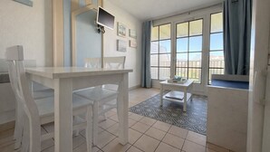 Ein Zimmer mit Esstisch, Stühlen, einem kleinen Beistelltisch, einem an der Wand montierten Fernseher und großen, mit Vorhängen versehenen Fenstern.
