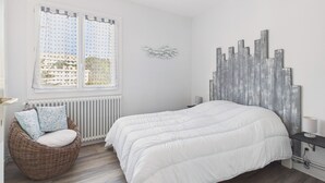 Ein Schlafzimmer mit einem großen Bett, einem Korbsessel mit Kissen, einem Heizkörper und einem Fenster mit Vorhängen.