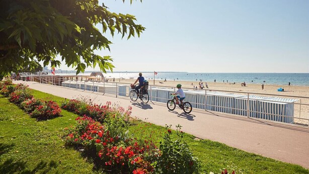 Ein Weg mit Geländer, im Hintergrund ein Strand und Menschen in Rollstühlen sowie auf Fahrrädern.