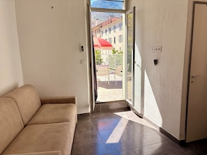 Een kamer met een beige bank, een schuifdeur naar een terras buiten met een rode parasol, en een uitzicht op gebouwen in de verte.