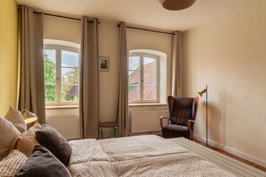 A bedroom with a bed, two windows, a chair, a lamp, and a radiator. - Alter Lindenhof Gnissau – Spacious Apartment in Historic Manor House (Ahrensbök)