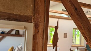 Interior rústic de fusta amb bigues descobertes, una làmpada penjant i vista cap a una zona de menjador per una finestra.