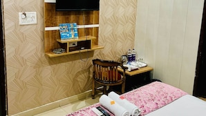 A hotel room with a bed, a TV mounted on the wall, a desk with a chair, and a shelf with items. - Hotel RAJ (Haridwar)