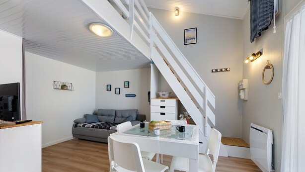 A modern interior with a staircase, a dining area with a glass table, white chairs, a grey sofa, and a kitchen area with a TV. - La Pause Iodée – Close to Beaches and Town Center (La Tranche-sur-Mer)