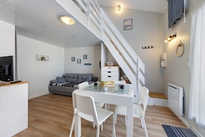 Un intérieur moderne avec un escalier, un coin repas doté d’une table en verre, de chaises blanches, d’un canapé gris et un espace cuisine avec un téléviseur.