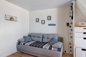 A grey sofa with striped throw, a wall-mounted shelf with a plant, and a sign indicating 'PLAGE CLEMENCEAU'. - La Pause Iodée – Close to Beaches and Town Center (La Tranche-sur-Mer)