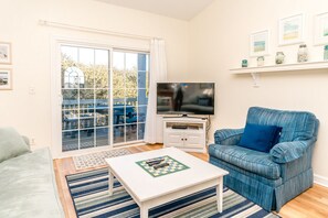 Ein Wohnzimmer mit Fernseher, einem blauen Sessel, einem weißen Couchtisch und einer Glastür, die zu einem Außenbereich mit blauem Tisch und Stühlen führt.