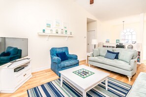 Ein Wohnzimmer mit Fernseher, blauem Sessel, hellgrauem Sofa, weißem Couchtisch und einem Streifenmuster-Teppich.