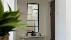 A hallway with a white wall, a wooden door, a window with a grid pattern, and a shelf with decorative items.
