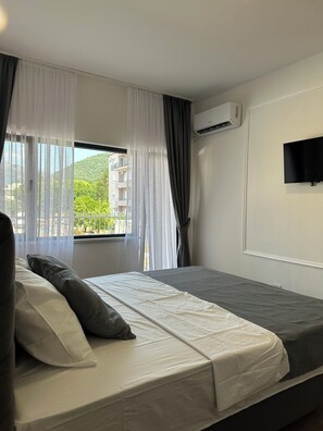Ein Schlafzimmer mit Bett, einem Fenster mit Vorhängen, einer Klimaanlage und einem an der Wand befestigten Fernseher.
