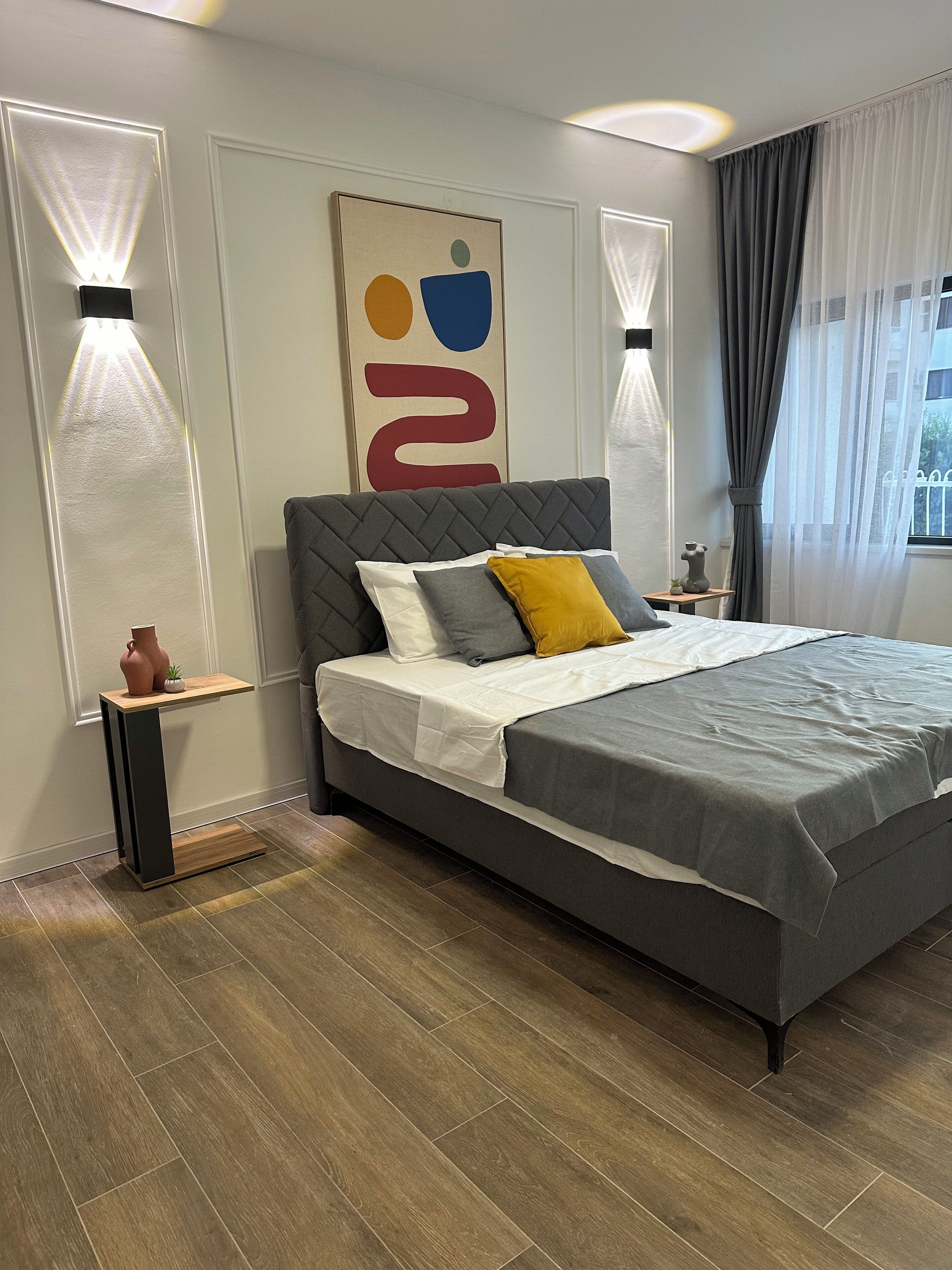 A modern bedroom with a large bed, a bedside table, a wall-mounted artwork, and a window with curtains.