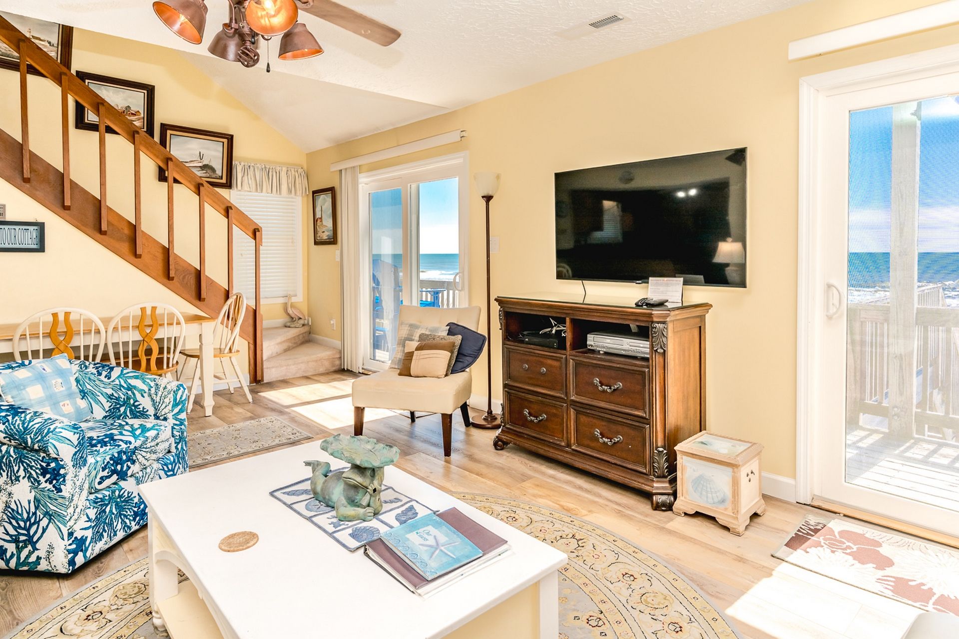 A spacious living room with a large TV, wooden staircase, and a view of the ocean.