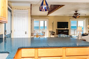 A kitchen with a blue countertop, wooden cabinets, a dining area with chairs, a television, and a view of the ocean. - Whittington - Oceanfront 3-Bedroom, perfect for family getaways! (North Topsail Beach)
