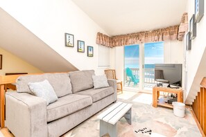 Ein Wohnzimmer mit einer Couch, einem Couchtisch und einem Fernseher auf einem Schrank. An den Wänden hängen Bilder in Rahmen und es gibt einen Balkon mit Blick.