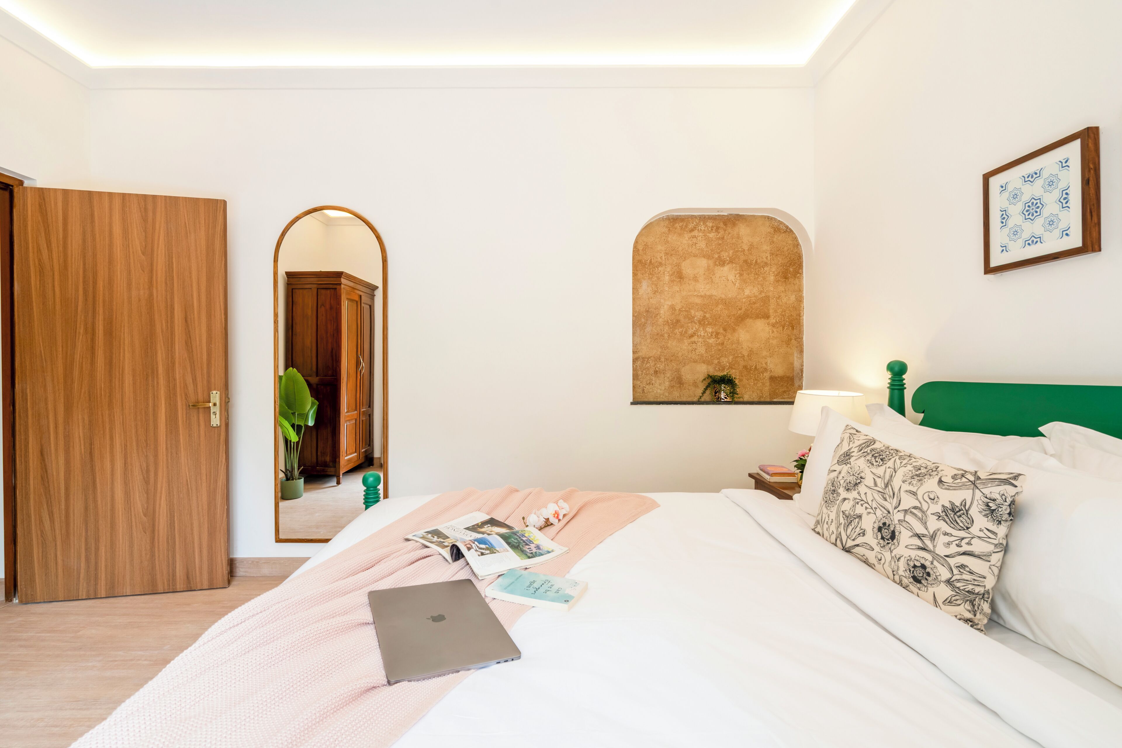 Una camera d'albergo con un letto, un portatile, uno specchio, una testiera verde e un quadro appeso al muro.