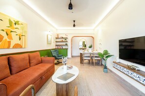 A living room with a sofa, a coffee table, a TV, a dining area with chairs, a bookshelf, and a mirror. - Staymaster Enchanto 2BR Apartment (Siolim)
