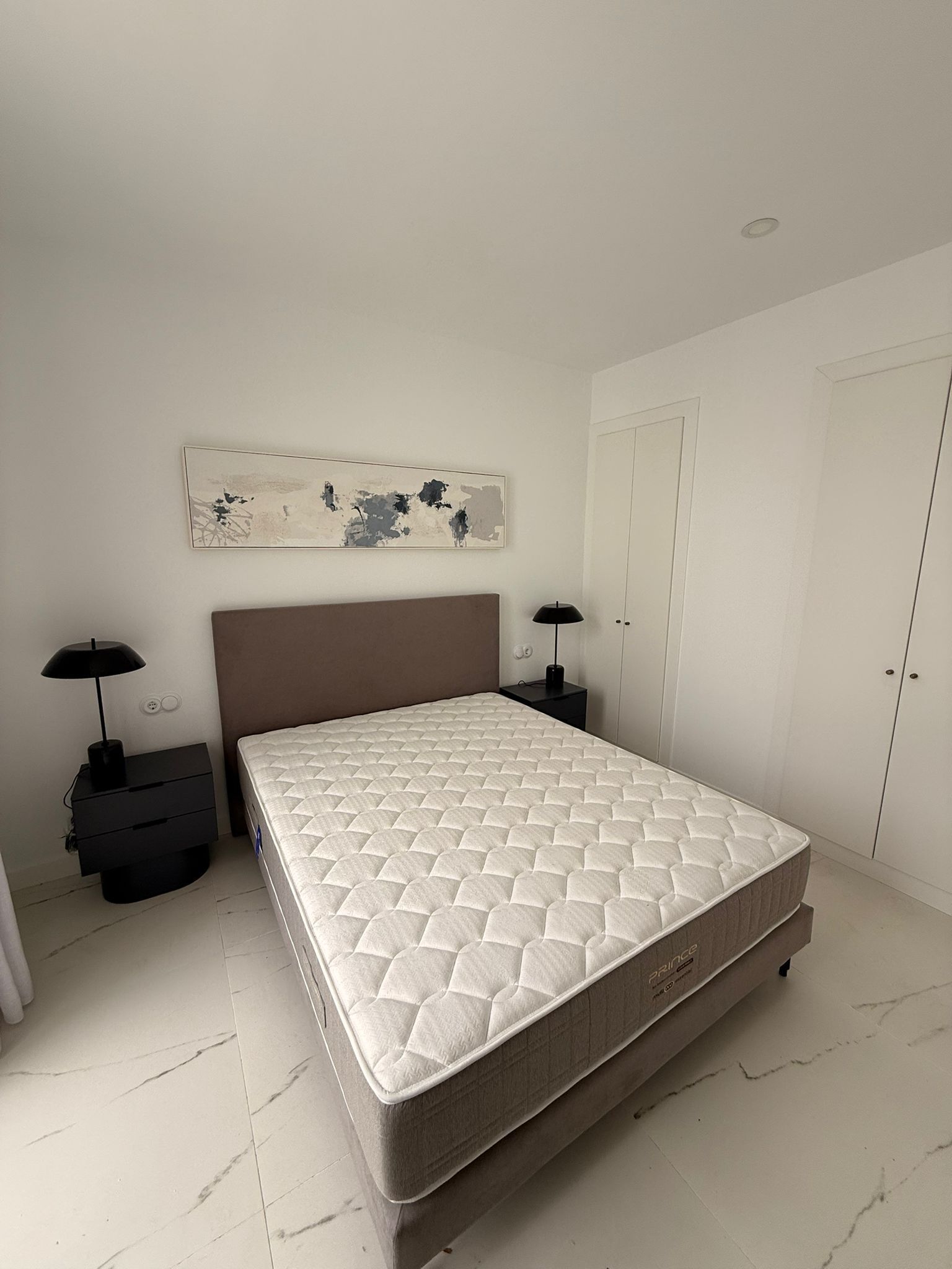 A bedroom with a queen-sized mattress, a headboard, a bedside table with a lamp, a wall-mounted abstract painting, and a wardrobe.