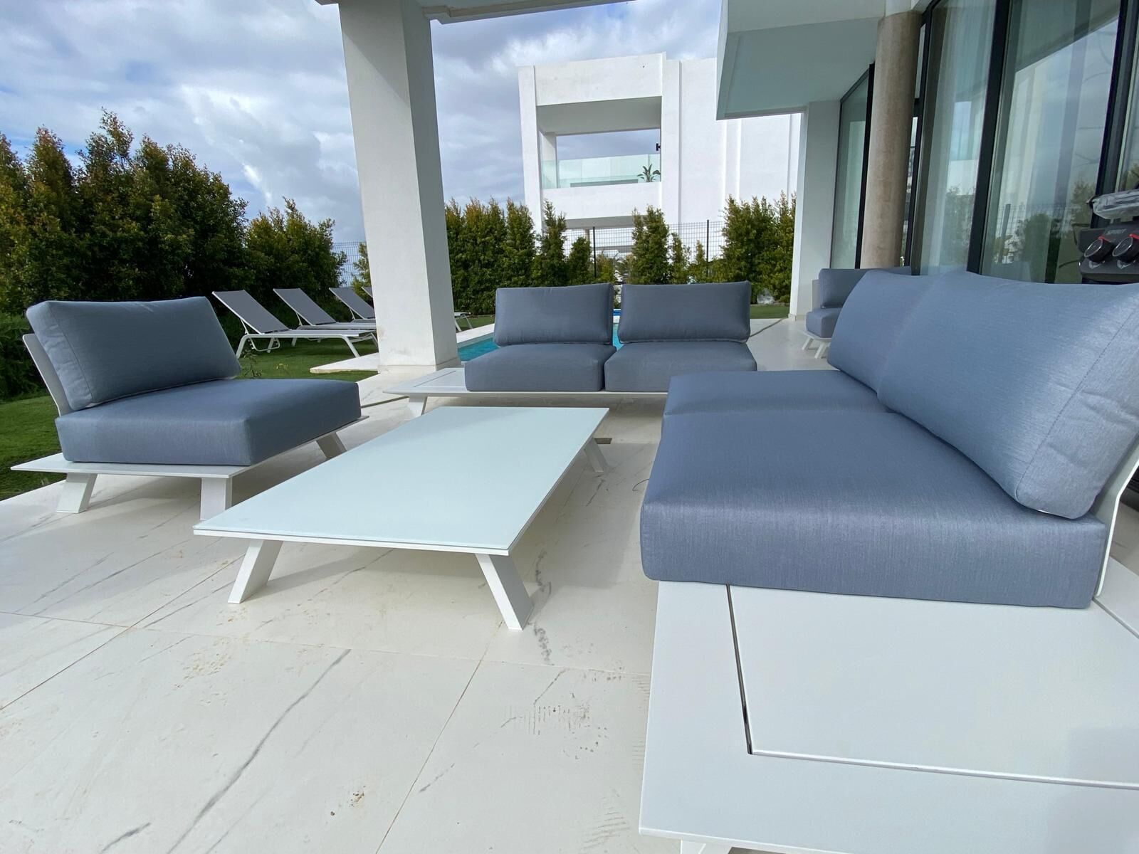 Un área de estar moderna al aire libre con mobiliario blanco y gris, una piscina y un edificio con grandes ventanales al fondo.