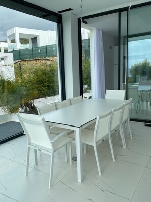 A modern dining area with a white table and chairs, large windows, and a view of the surrounding landscape. - Villa Harmonia 22, SOTOGRANDE (San Roque)