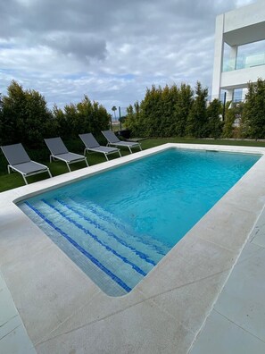 A rectangular swimming pool with a tiled edge, surrounded by a concrete deck. There are four lounge chairs aligned along the pool's edge. - Villa Harmonia 22, SOTOGRANDE (San Roque)